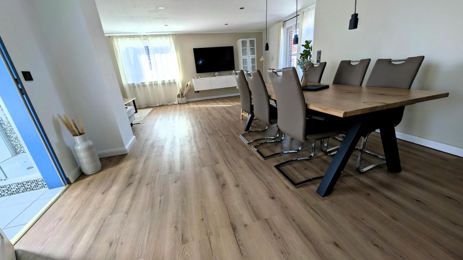 Essbereich mit Holztisch und Stühlen. Im Hintergrund Wohnzimmer mit Fernseher und Fenstern.