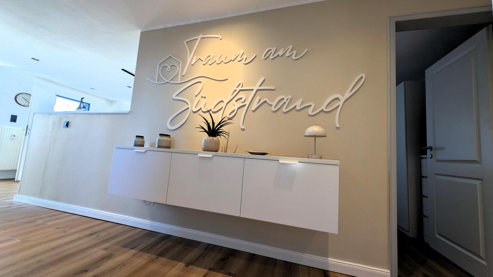 Wand mit Schriftzug Traum am Südsttrand und weiße Sideboard mit Pflanze und Lampe.