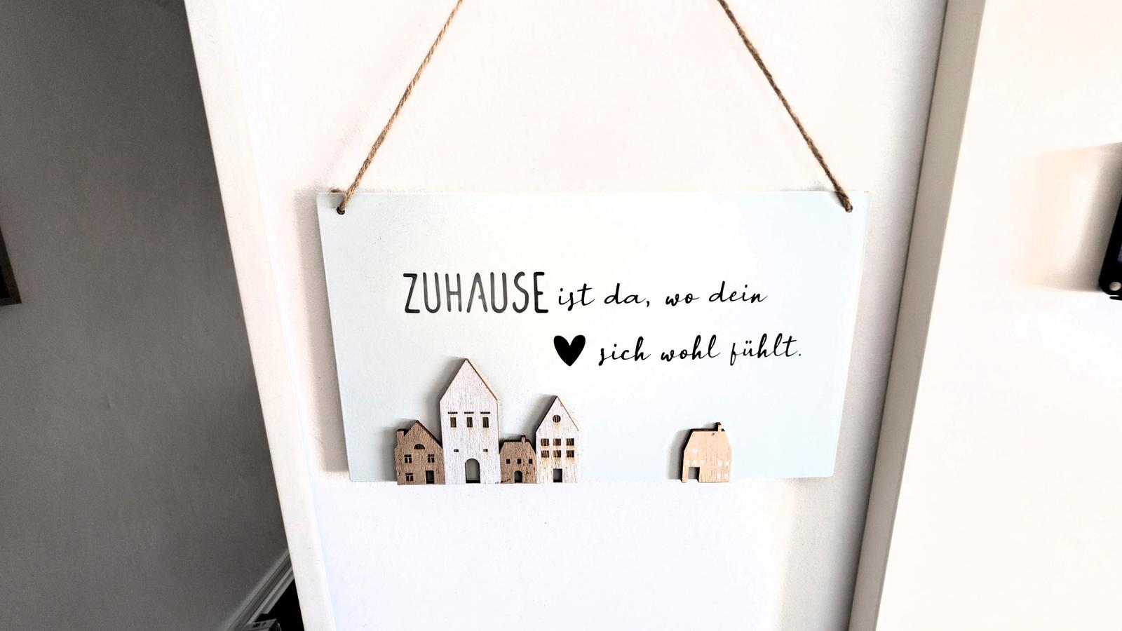 Hängendes Schild mit Text und Holzhäusern an Wand