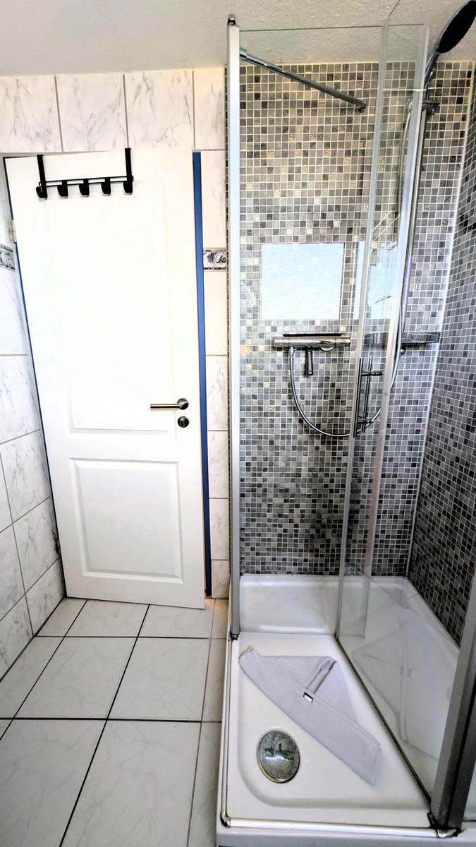 Bad mit Dusche und Fliesenwand. Tür und Bodenfliesen sind weiß.