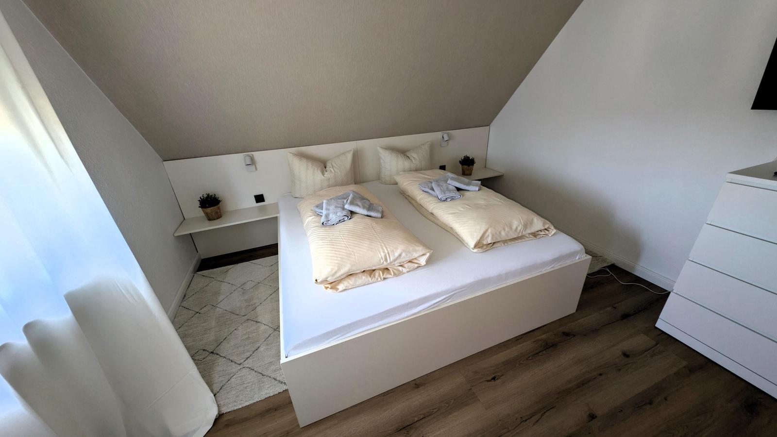 Schlafzimmer mit Doppelbett, Holzboden und Fenster. Weiße Wand und Bettgestell.