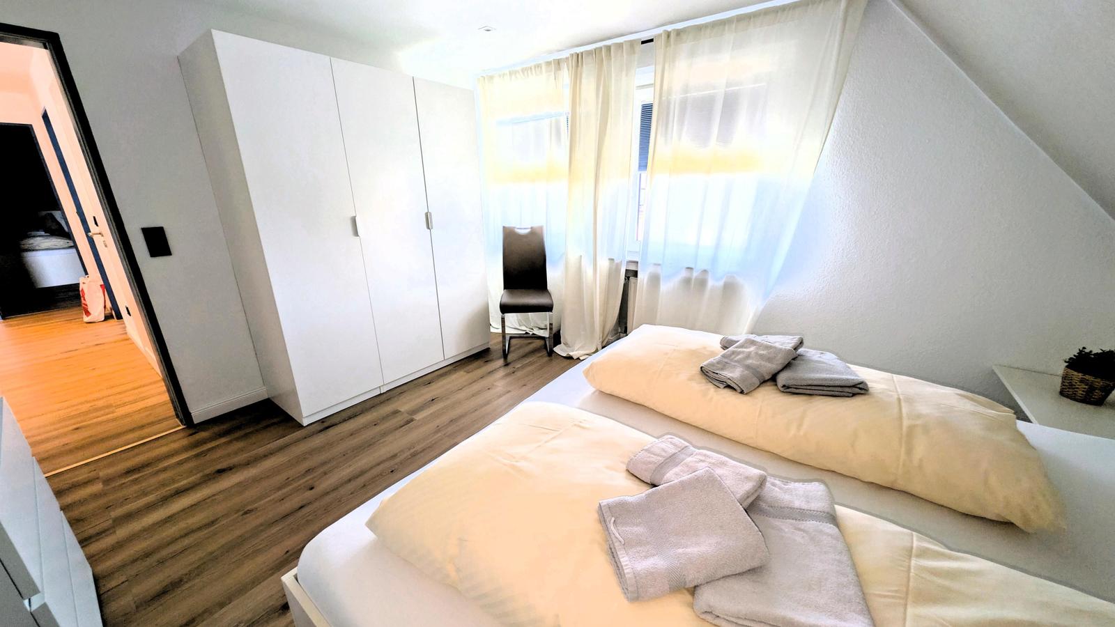Schlafzimmer mit Doppelbett, weißen Schränken und Holzboden. Fenster mit weißen Vorhängen.