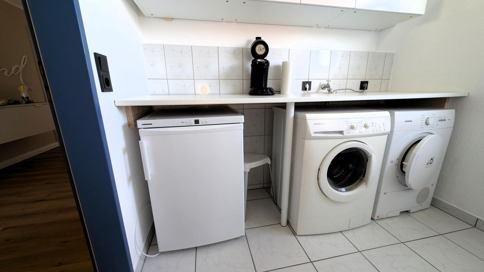 Waschmaschine, Trockner und Kühlschrank in einem hellen Raum mit Fliesenboden und Arbeitsplatte.