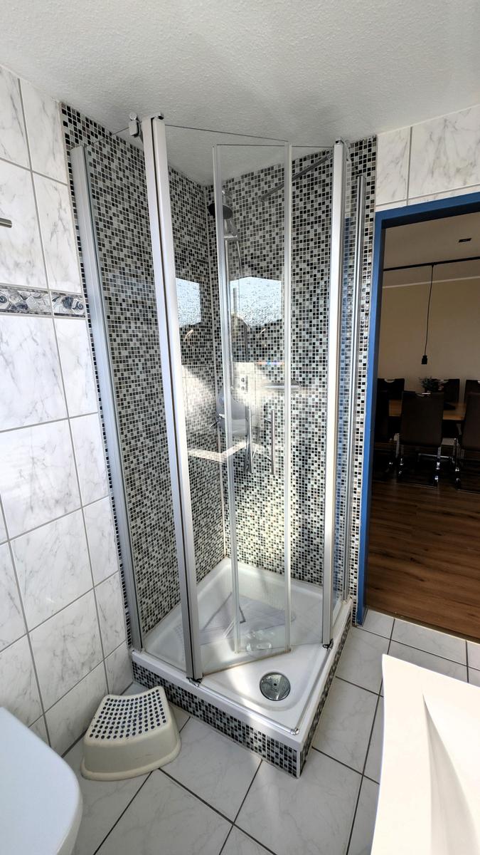 Dusche mit Glaswand und Mosaikfliesen, weiße Fliesen und Waschbecken sichtbar.