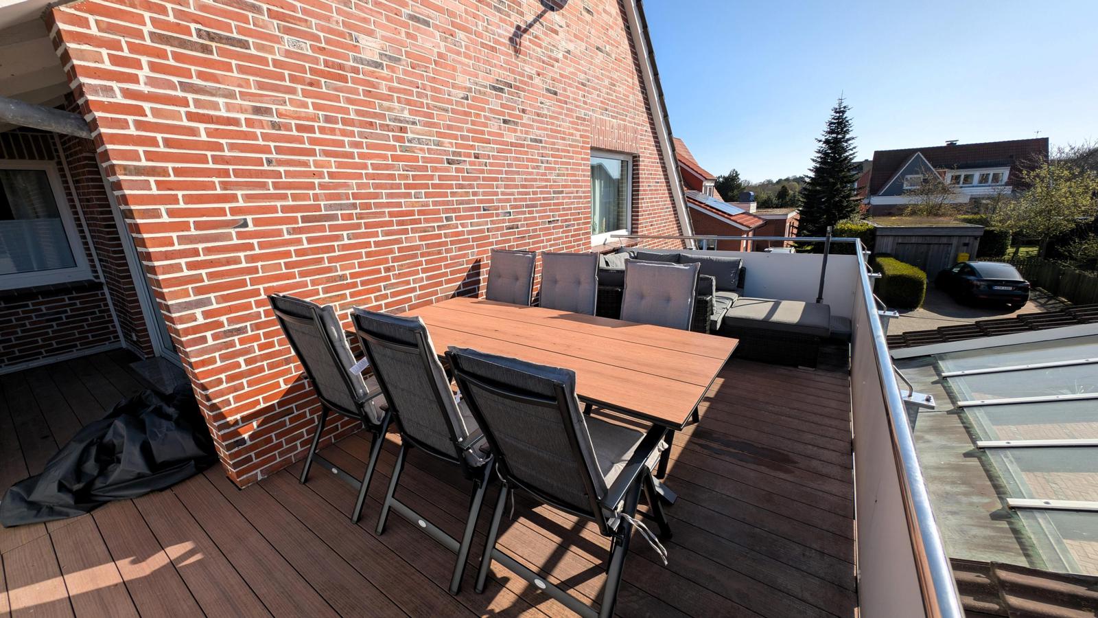 Terrasse mit Holztisch, Stühlen und Sitzgruppe. Hintergrund: Hauswand und Nachbarhäuser.