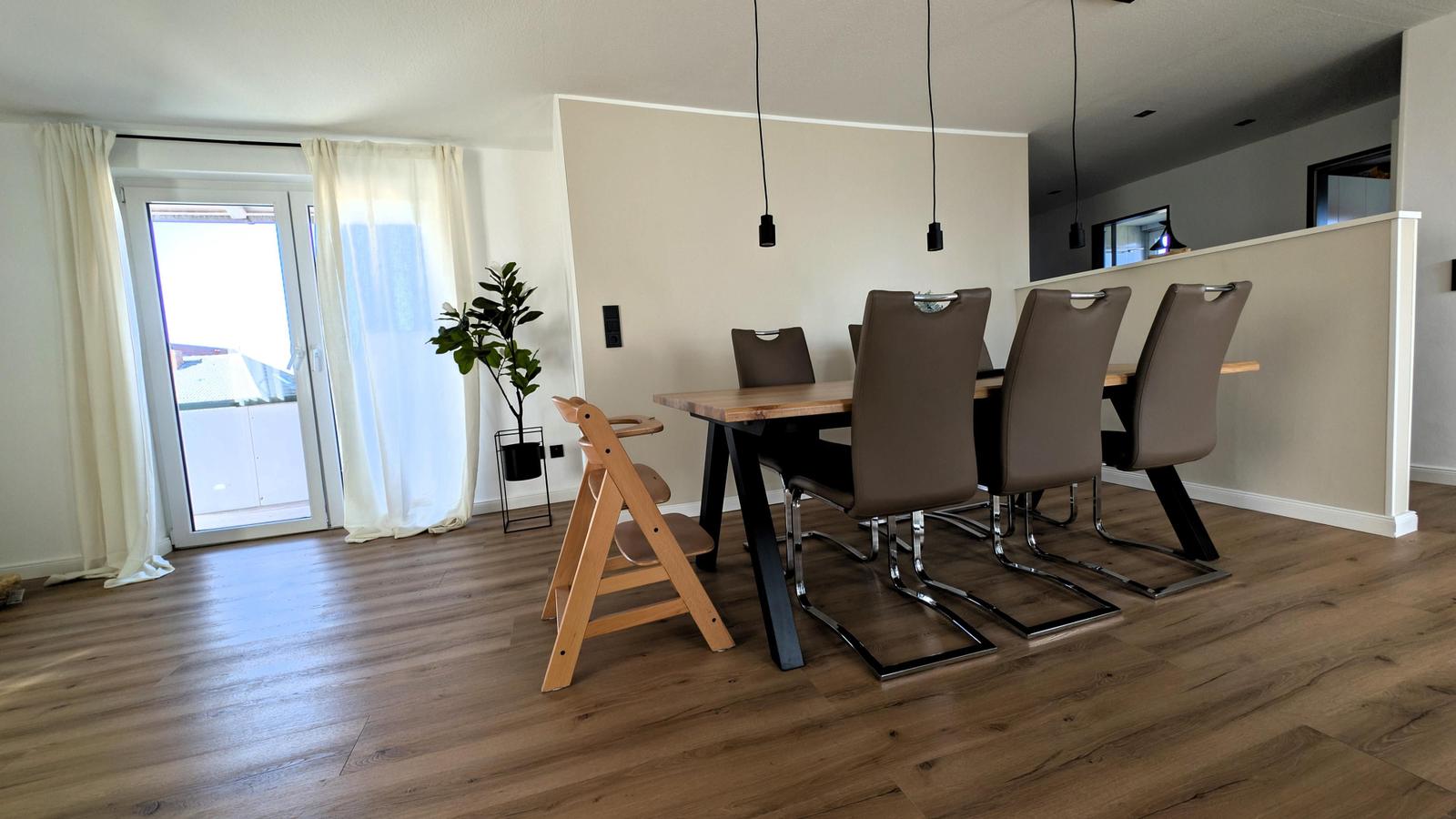 Esszimmer mit Holztisch, braunen Stühlen und Kinderschreibtisch. Durchgang zur Küche und Balkon mit Vorhängen.