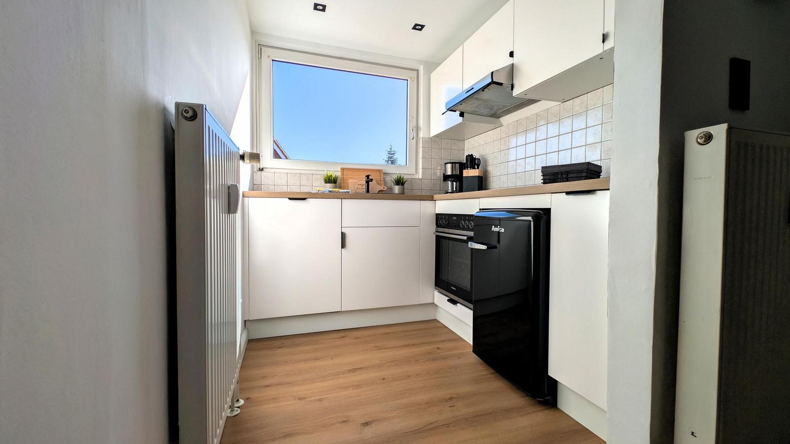 Küche mit weißen Schränken, Holzboden und Fenster. Schwarz-weiße Geräte und Küchenzeile.