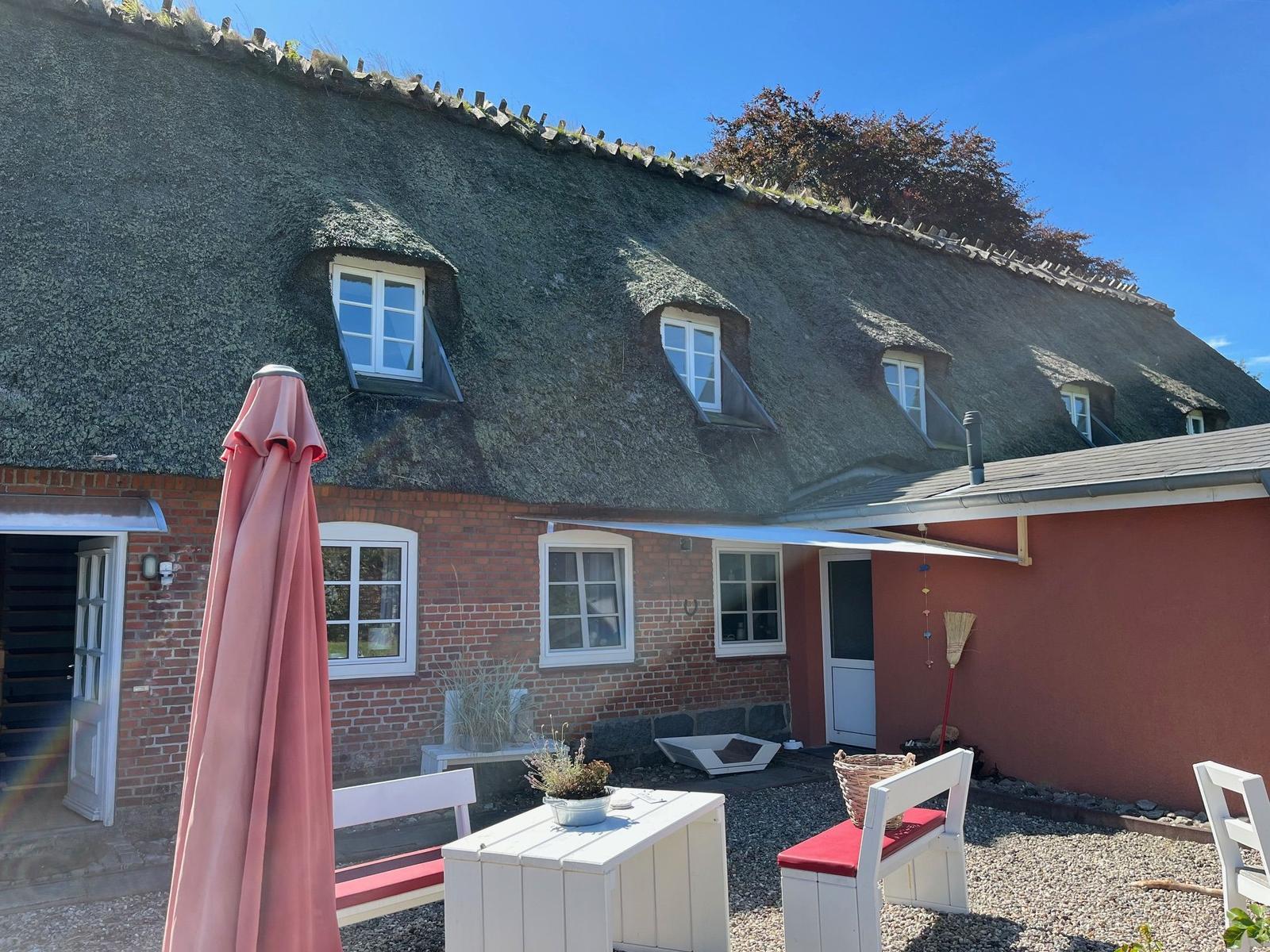 House with thatched roof, terrace, and garden furniture under blue sky.