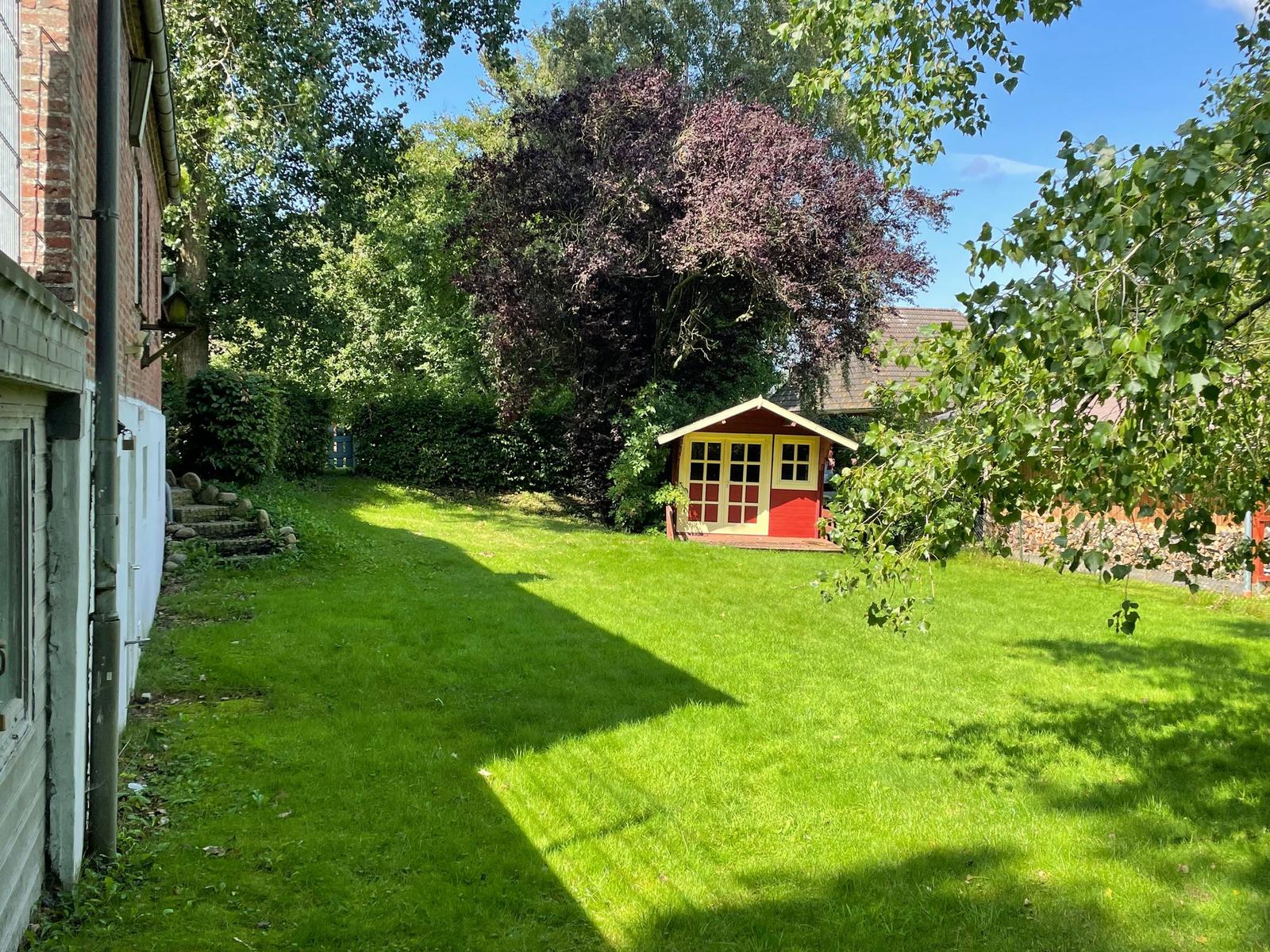 Hinterhof mit grünem Rasen, kleinem rotem Holzhaus und Bäumen.