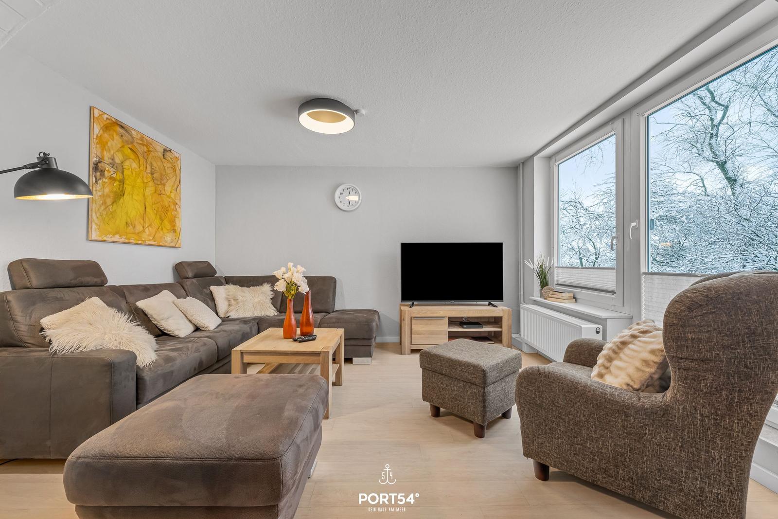 Wohnzimmer mit Sofa, Sessel und Fernseher. Große Fenster mit Blick auf schneebedeckte Bäume.