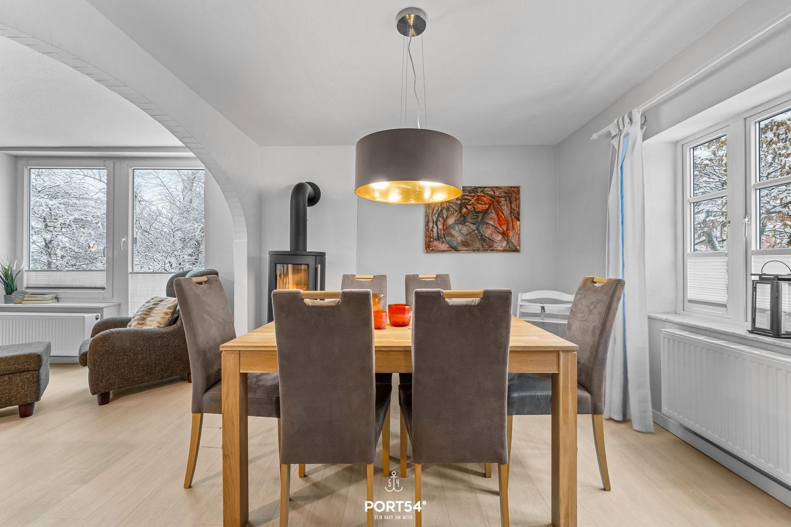 Esszimmer mit Holztisch, Stühlen und Kaminofen. Fenster zeigen schneebedeckte Bäume.