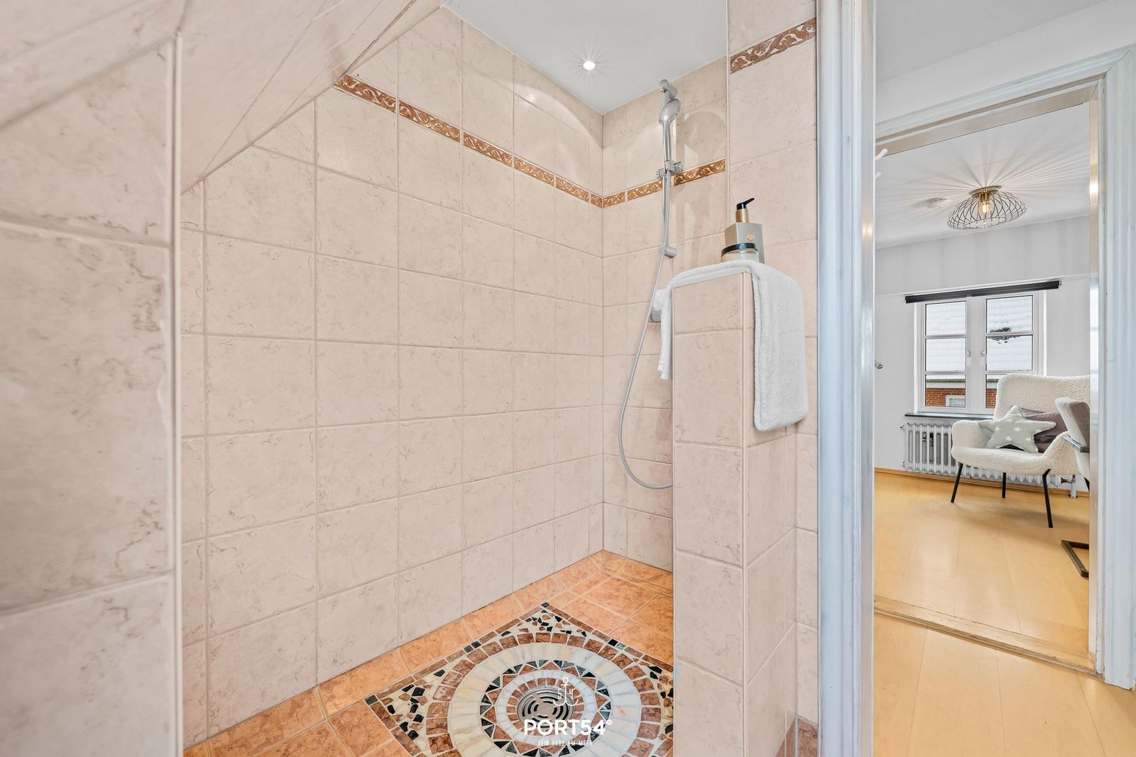 Duschkabine mit Mosaikboden und Handbrause. Im Hintergrund ist ein Wohnzimmer mit Sessel sichtbar.