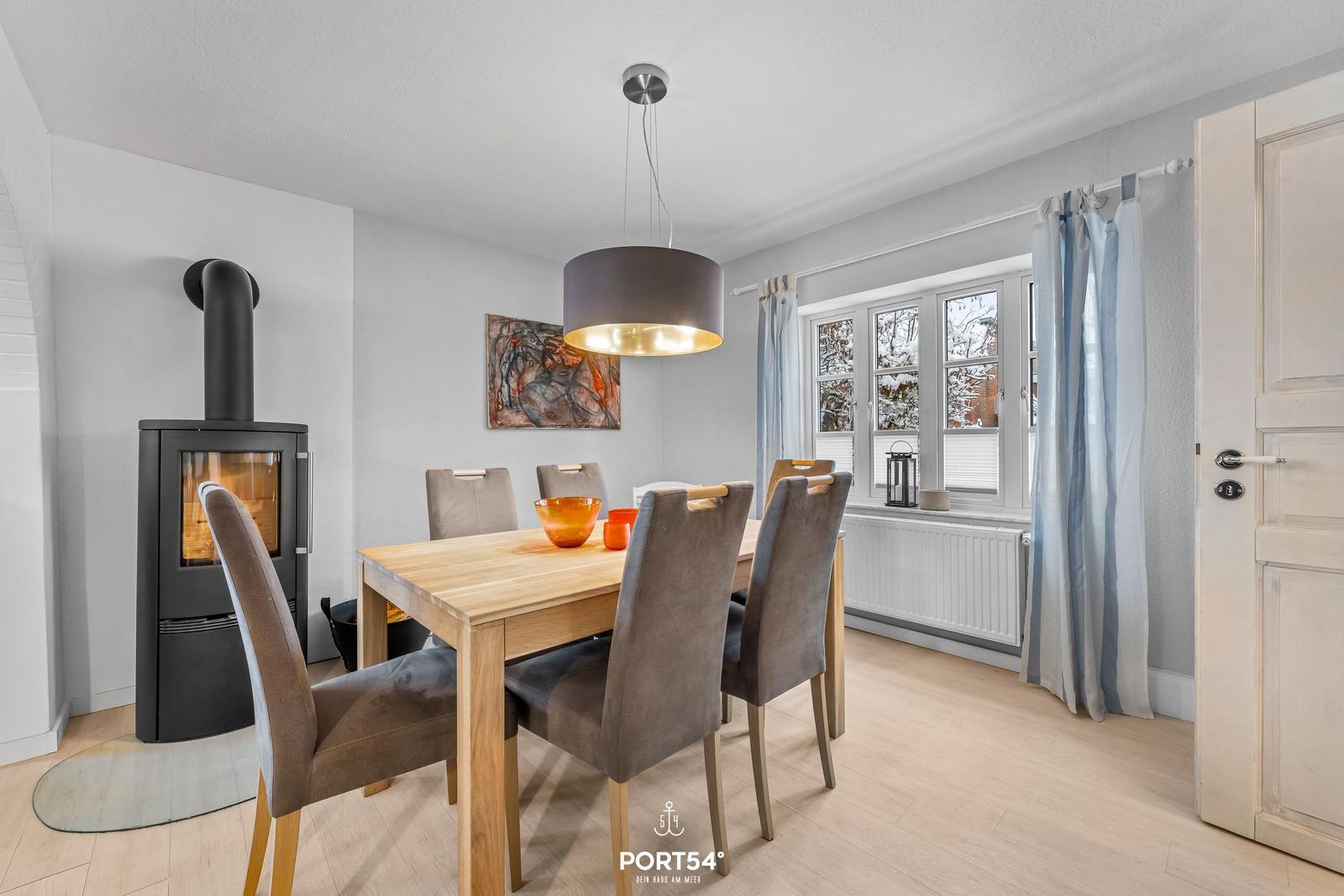 Esszimmer mit Holztisch, Stühlen und Kaminofen. Fenster mit Vorhängen.