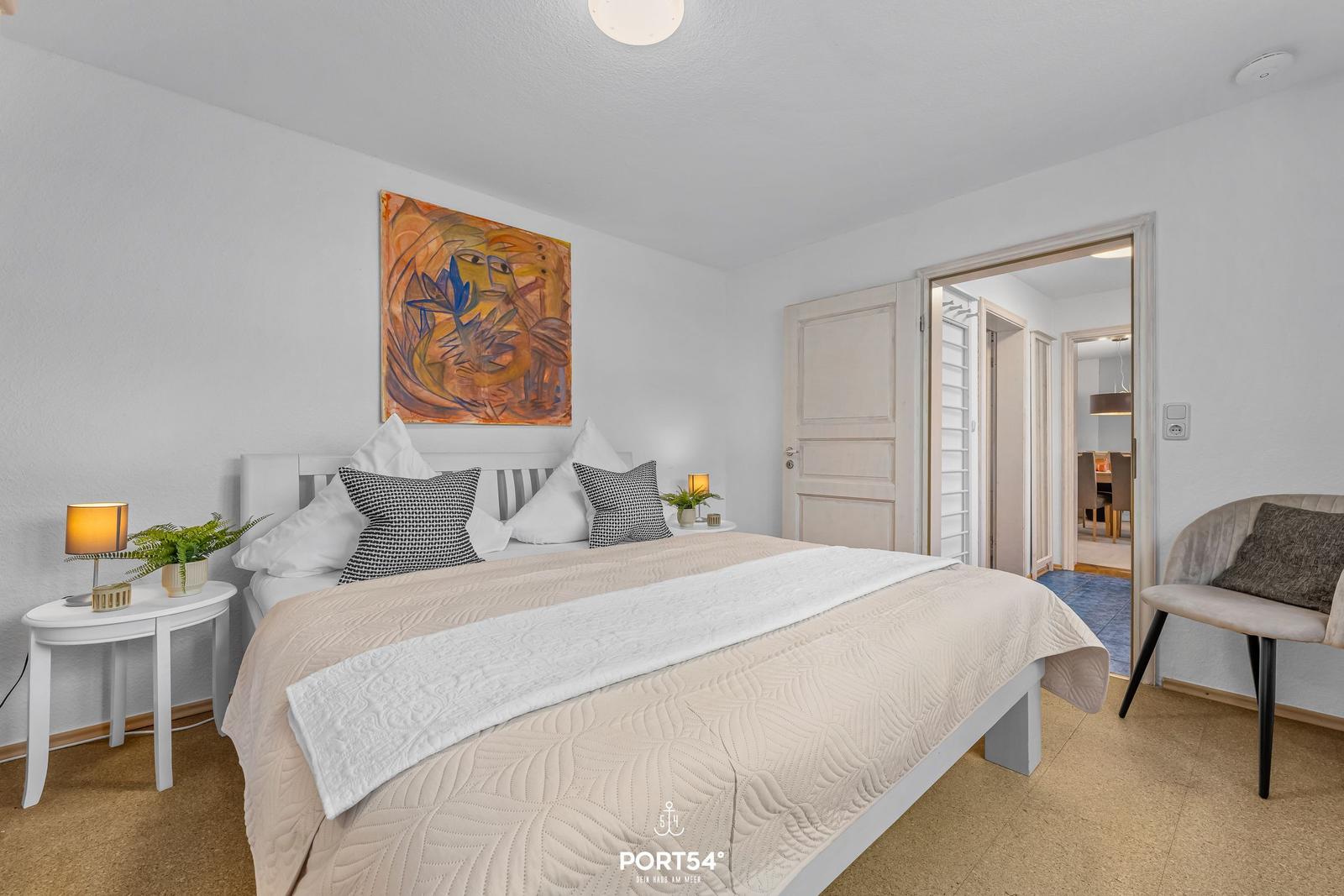 Schlafzimmer mit Bett, Nachttisch und Stuhl. Wandbild über dem Bett.