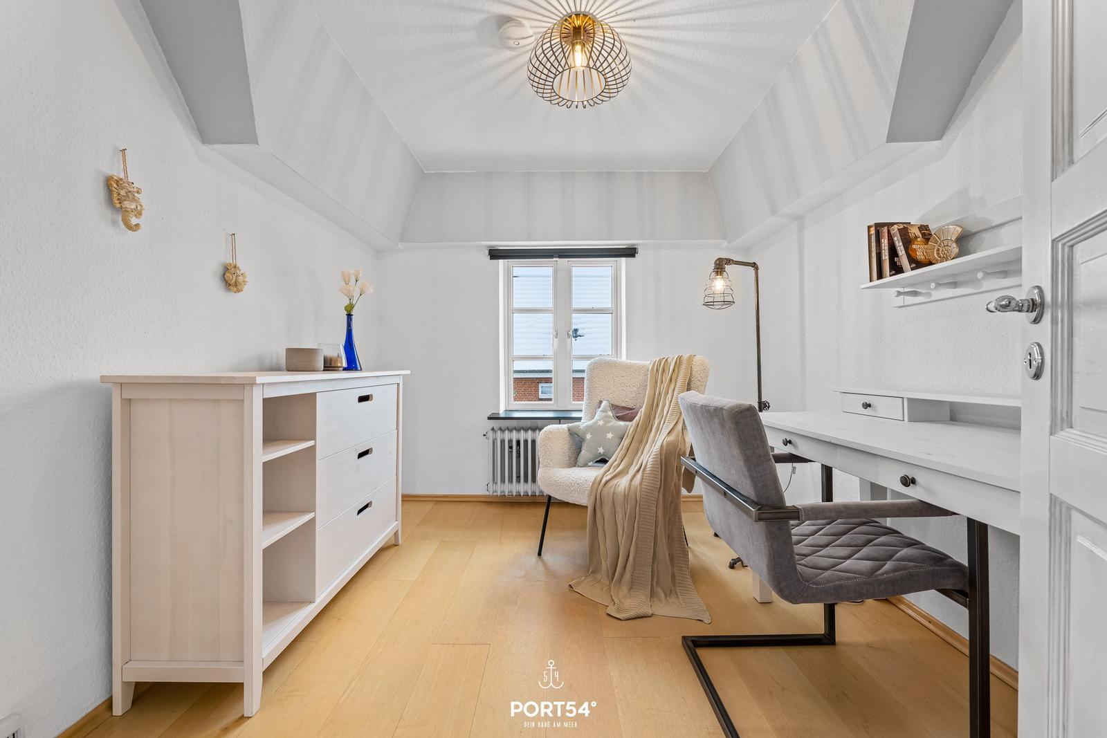 Schlafzimmer mit Schreibtisch, Sessel und Kleiderschrank. Holzboden und weiß gestrichene Wände.