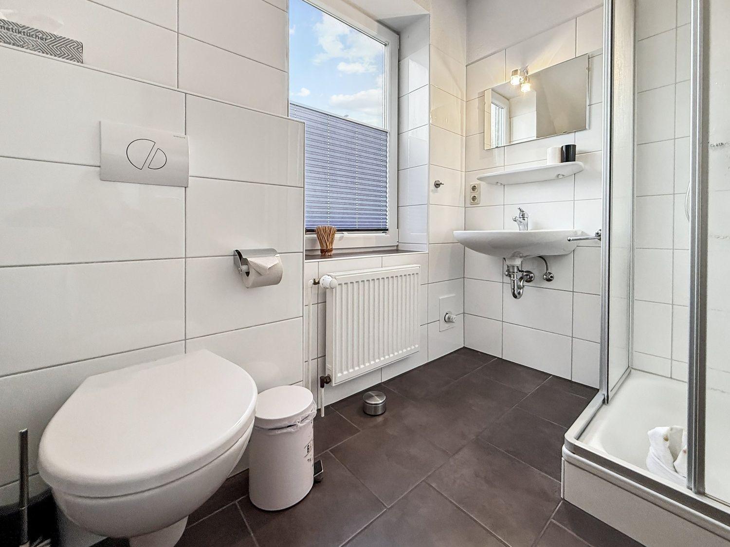 Badezimmer mit Toilette, Waschbecken und Dusche. Fenster mit Jalousie.