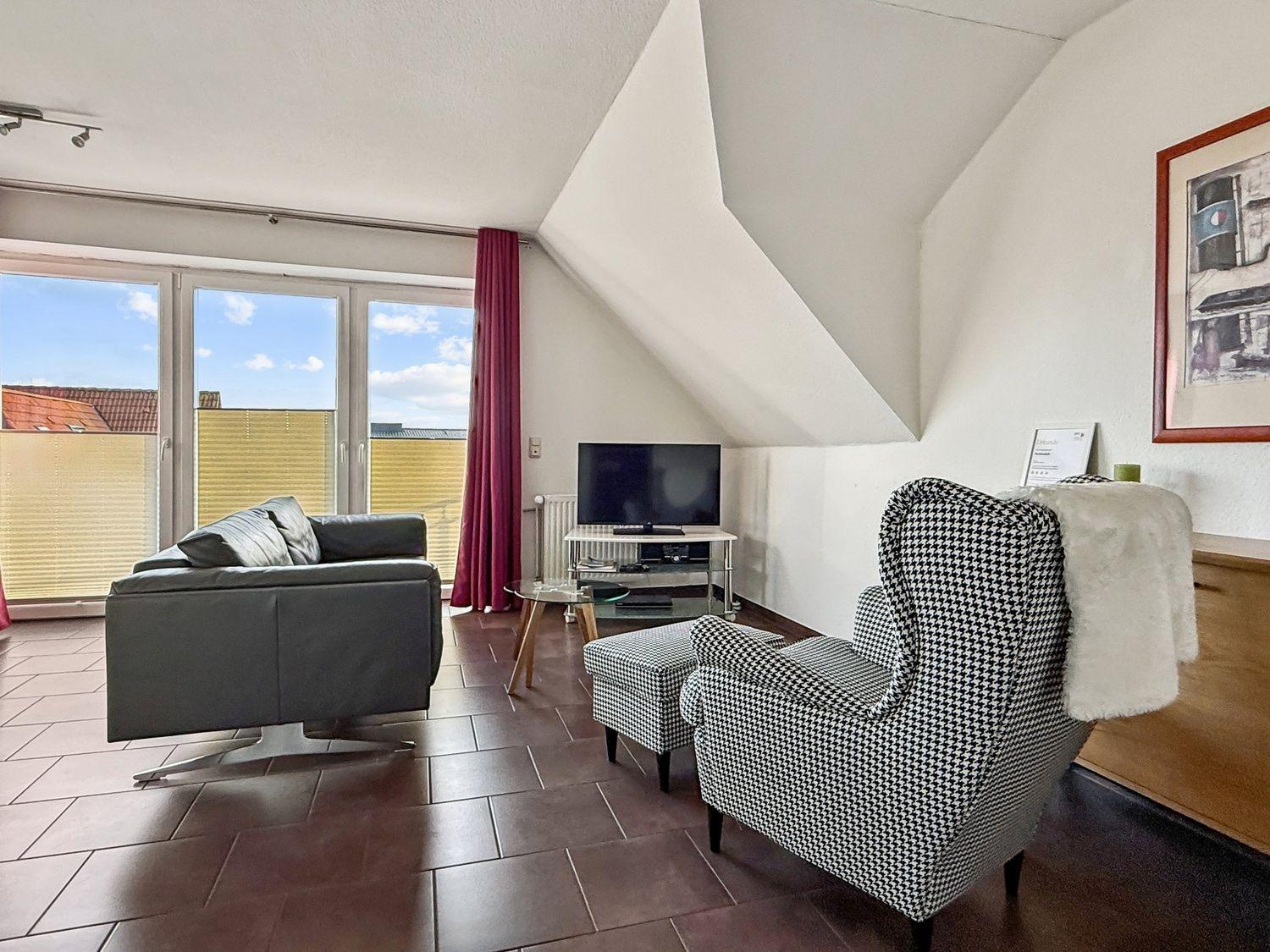 Wohnzimmer mit Sofa, Sessel und TV. Große Fenster und Dachschrägen.