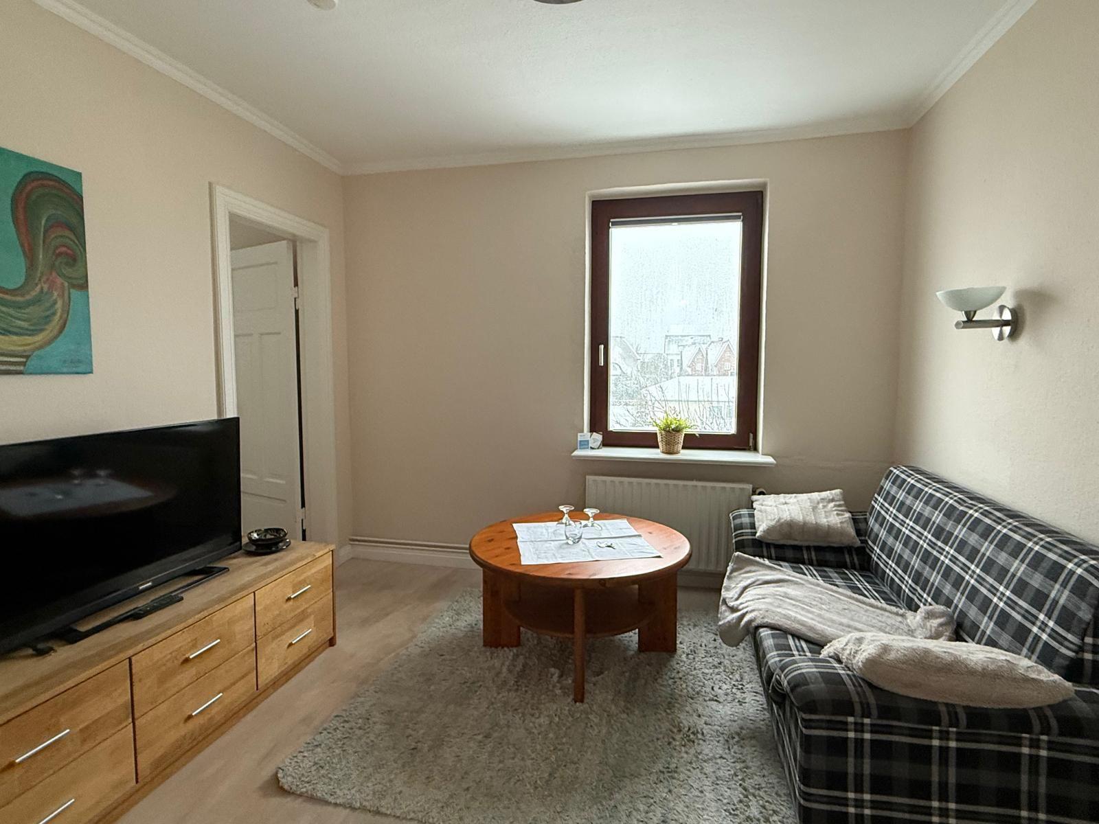 Wohnzimmer mit Sofa, Rundtisch und Fernseher. Fenster mit Blick auf Dächer.