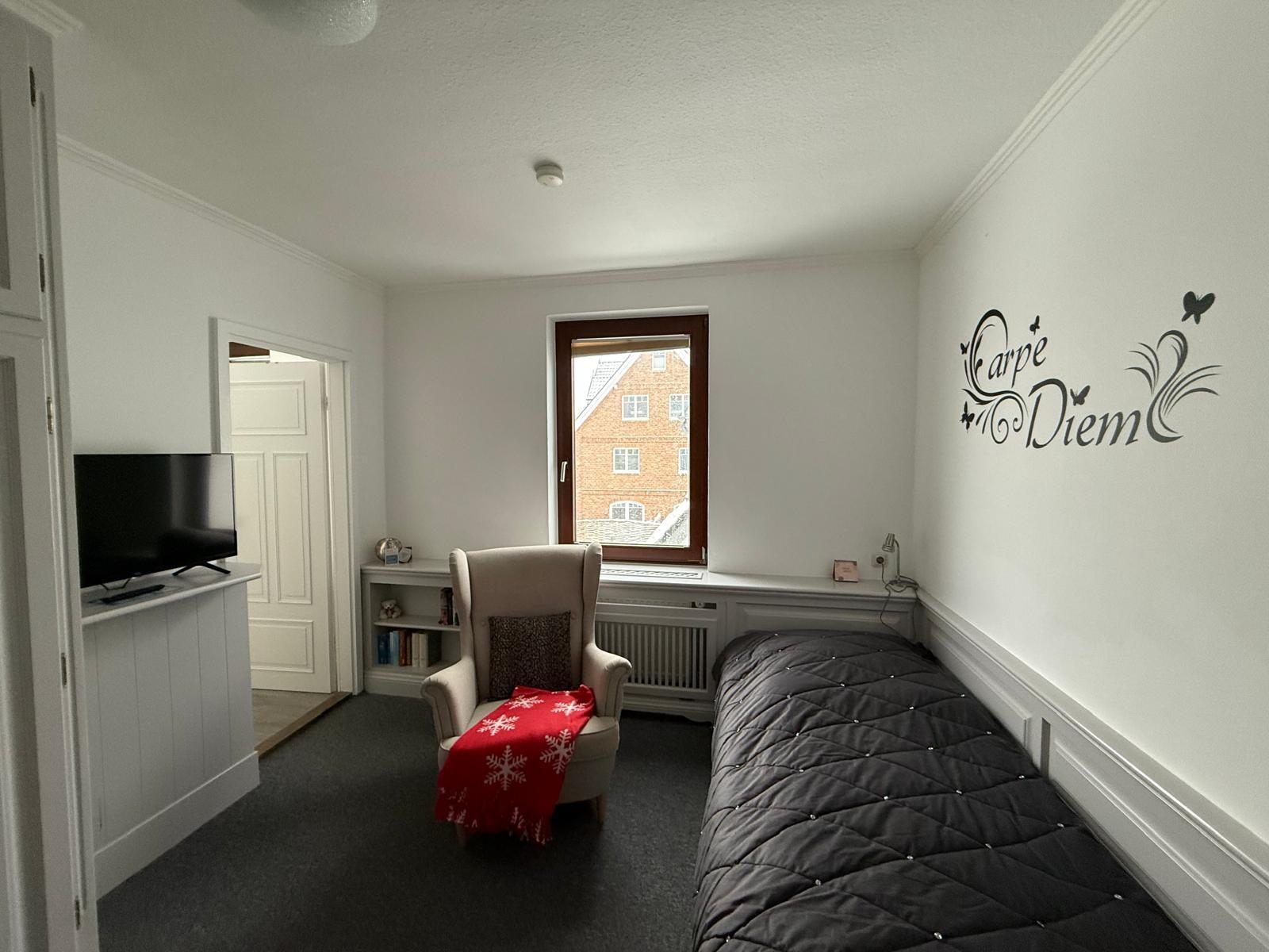 Schlafzimmer mit Bett, Sessel, Fernseher und Fenster mit Blick auf Haus.