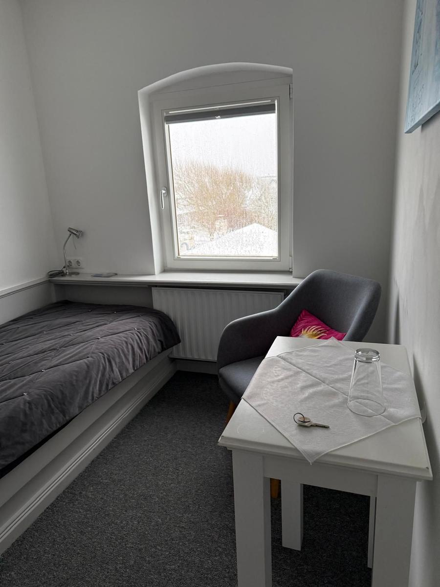 Zimmer mit Bett, Stuhl und Tisch. Fenster mit Blick auf Außenbereich.