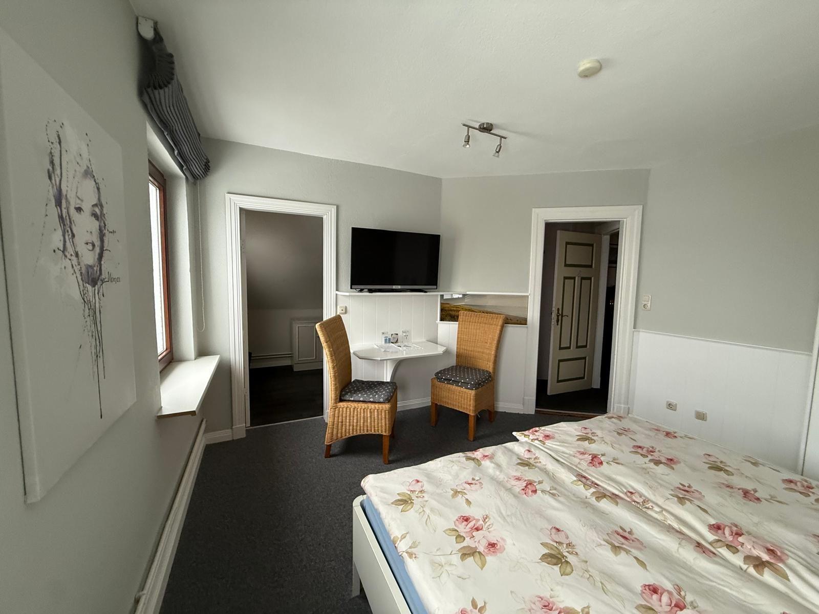 Schlafzimmer mit Bett, Fernseher und zwei Stühlen. Wandbild und Fenster sichtbar.