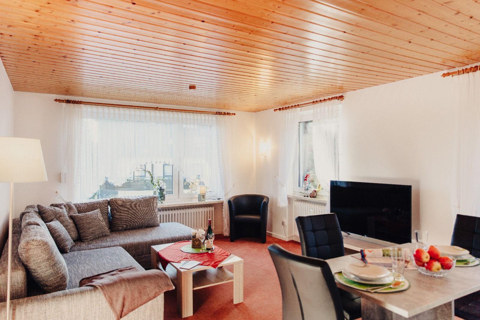 Wohnzimmer mit Sofa, Esstisch und Fernseher. Holzdecke und Fenster mit Vorhängen.