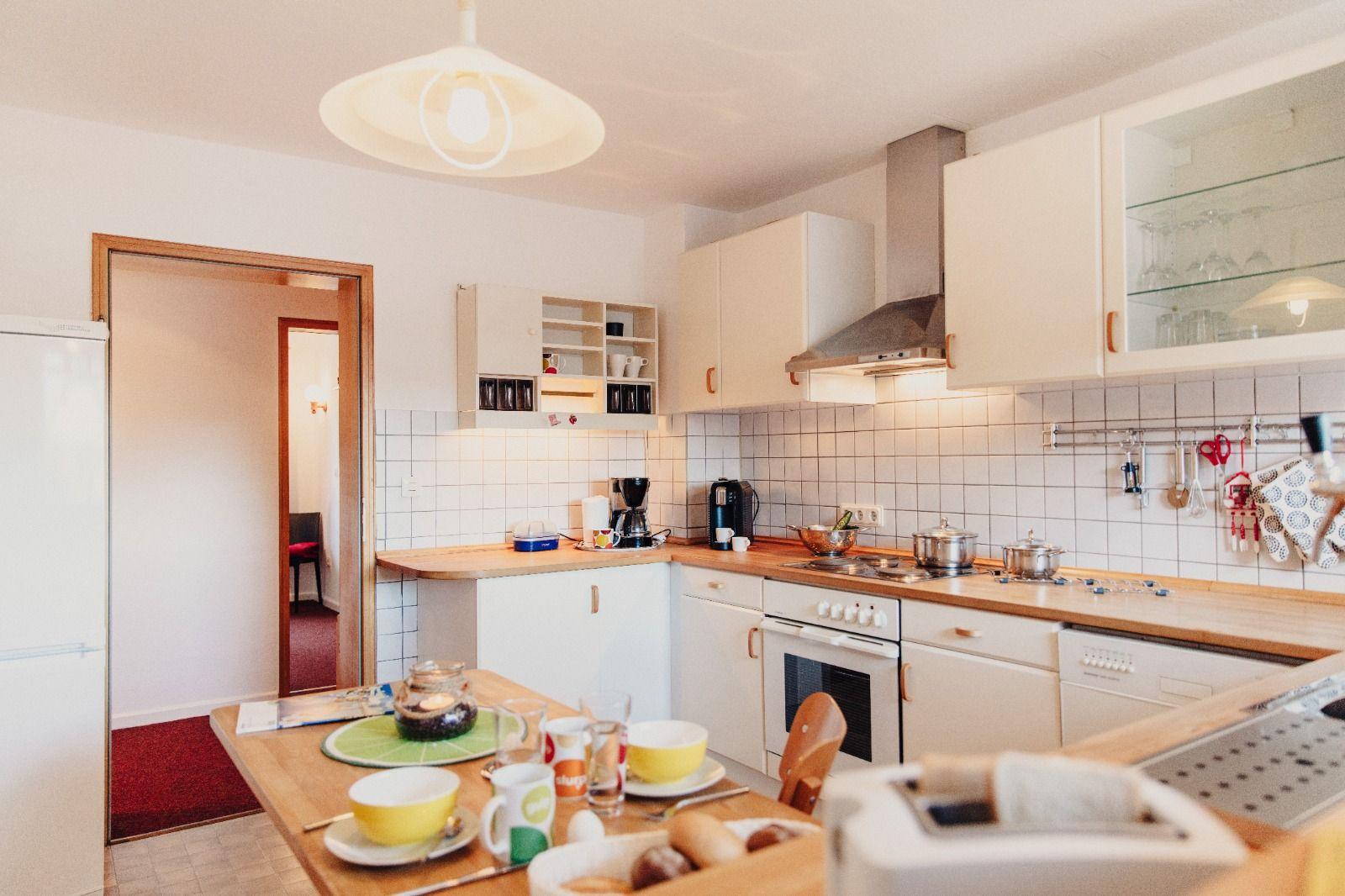 Küche mit Holzoberflächen, weißen Schränken und Küchenzeile. Esstisch mit Geschirr und Kaffeemaschine.