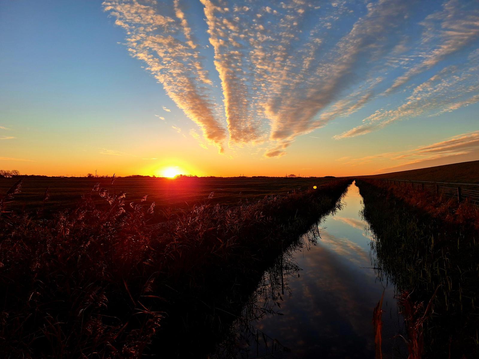 Sonnenuntergang über Fluss und Felder mit spiegelndem Himmel.