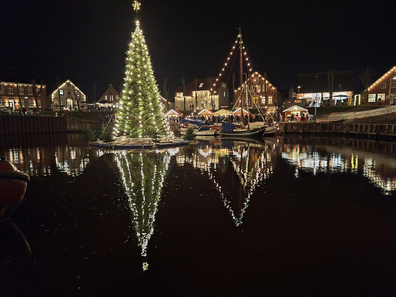 Weihnachtsbaum auf Wasser, beleuchtet, mit Reflexion. Hintergrund: beleuchtete Häuser und Marktstände.