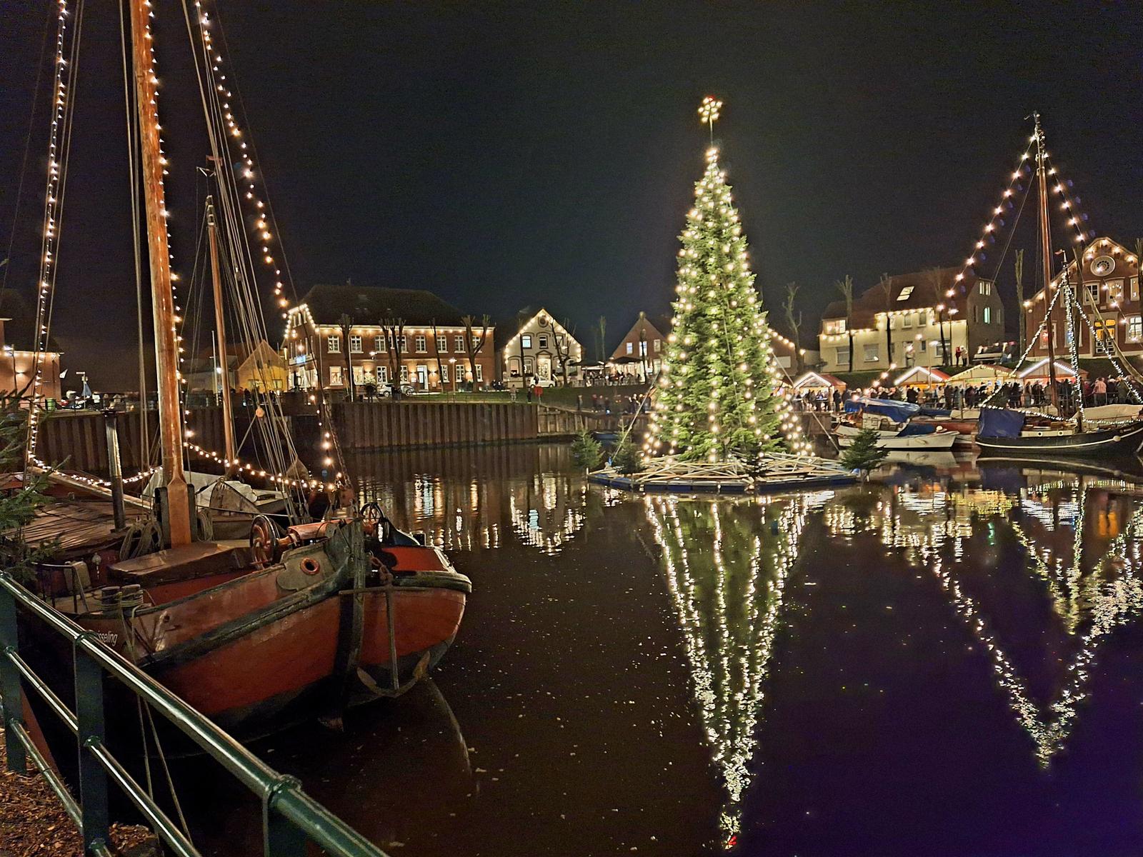 Weihnachtsbaum auf Wasser mit beleuchteten Booten und Häusern im Hintergrund.