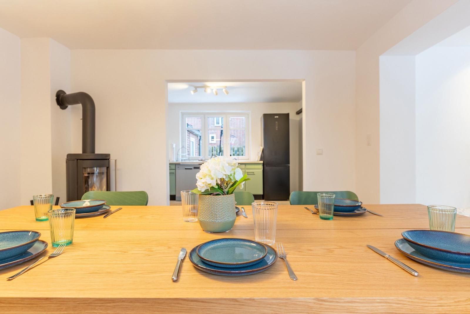 Esstisch mit blauen Tellern und grünen Stühlen. Im Hintergrund Küche und Kamin.