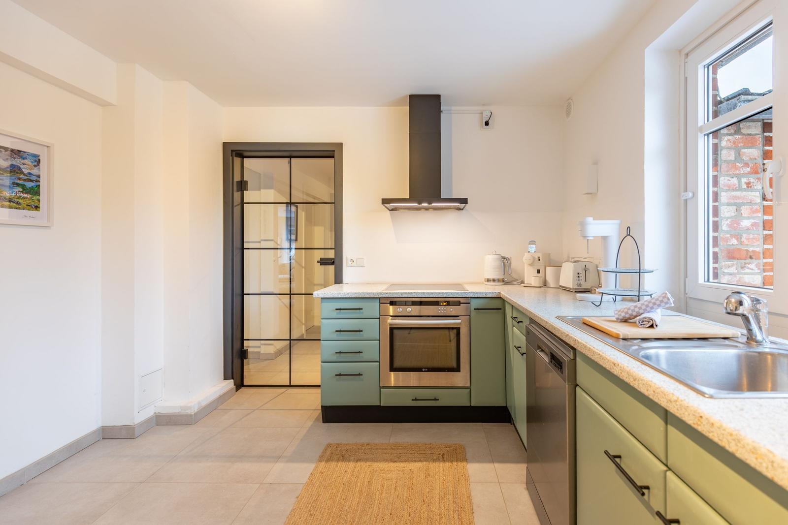 Küche mit grünen Schränken, Edelstahlgeräten und Fenster mit Blick auf Backsteinmauer.