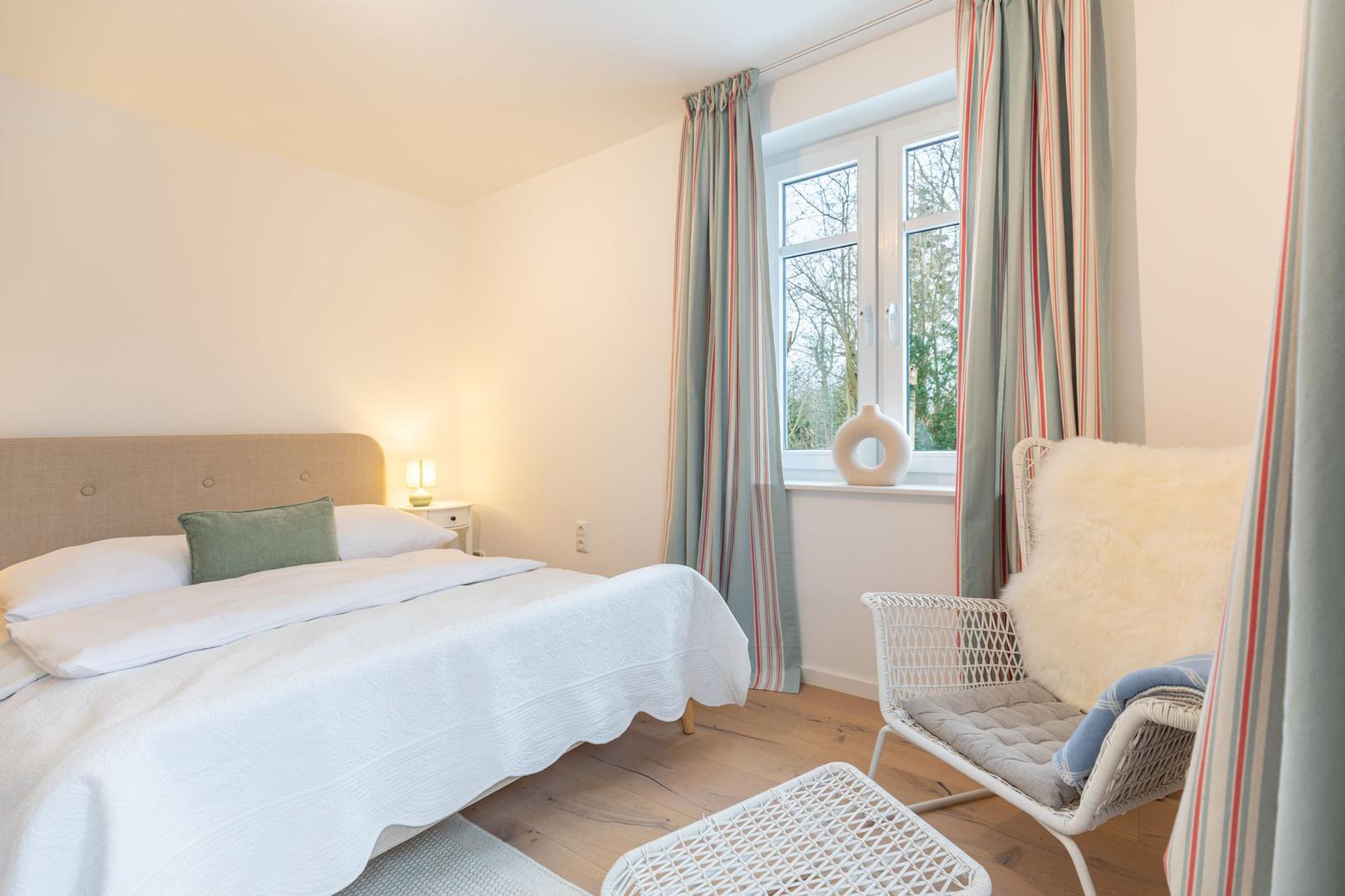 Schlafzimmer mit Bett, Sessel und Fenster mit Blick auf Außenbereich.