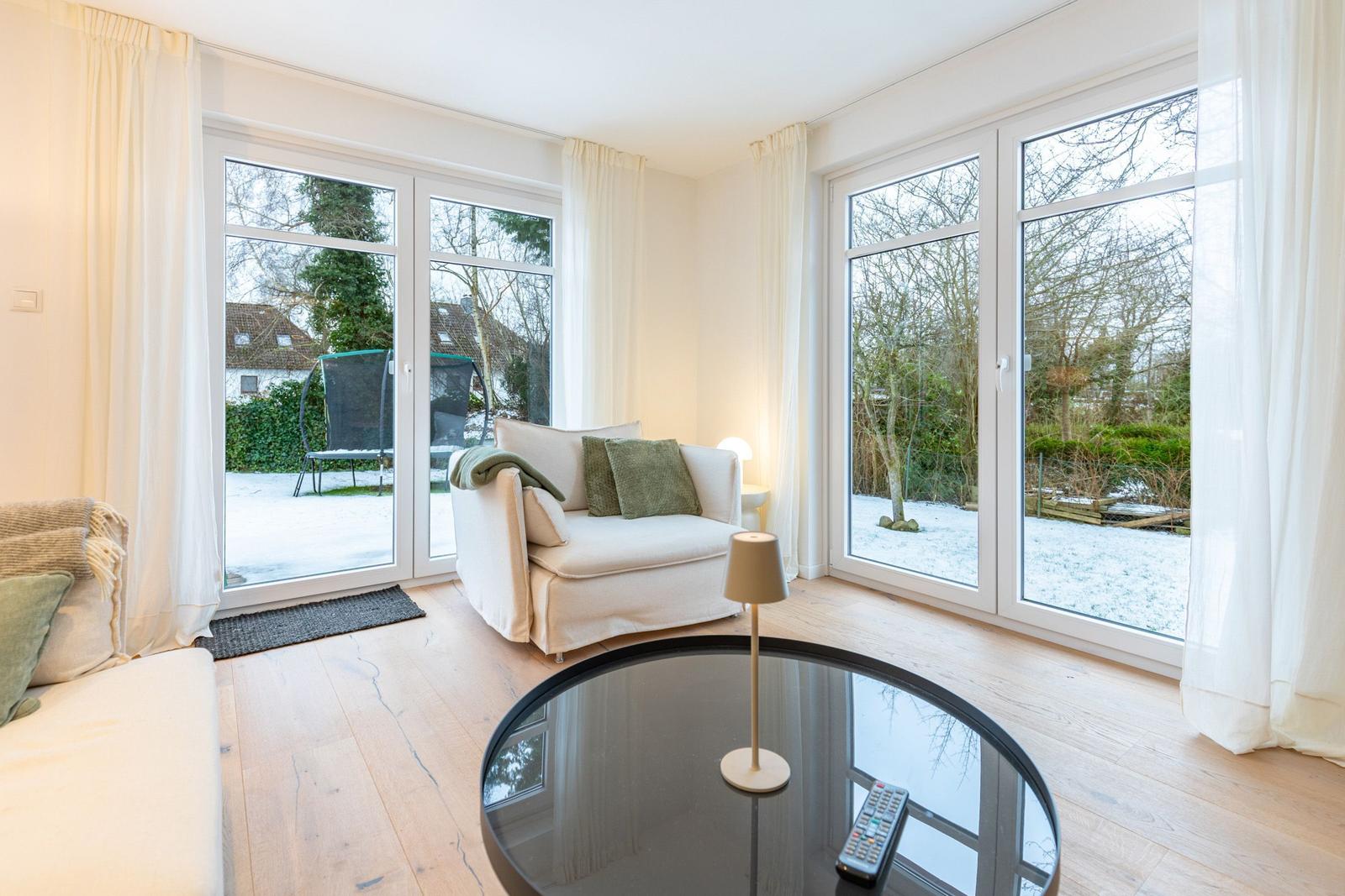 Wohnzimmer mit großem Fenster und Sitzgelegenheiten. Draußen Schnee und Garten.