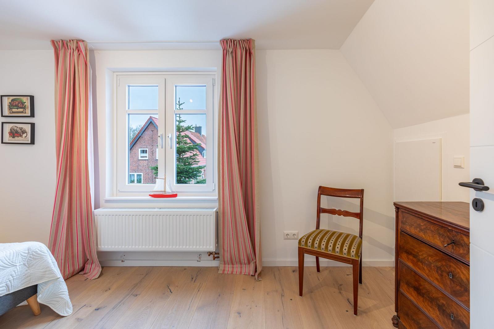 Schlafzimmer mit Fenster, Gardinen, Stuhl und Schrank.