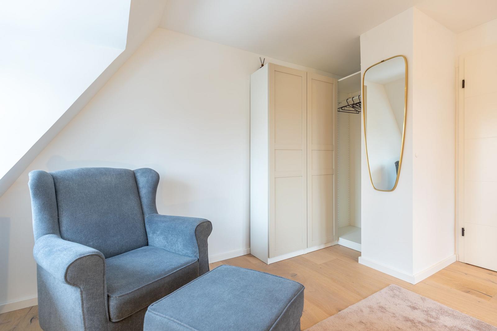 Ein Raum mit grauem Sessel, Kleiderschrank und Spiegel an der Wand.