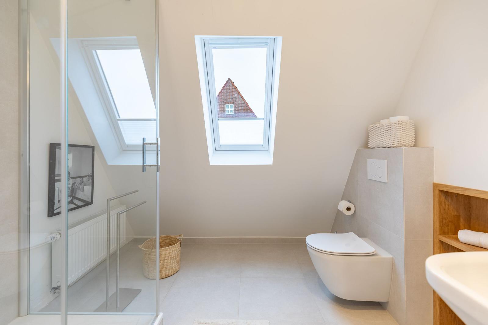 Badezimmer mit Dusche, WC und Fenster. Holzregal und Korb sind sichtbar.