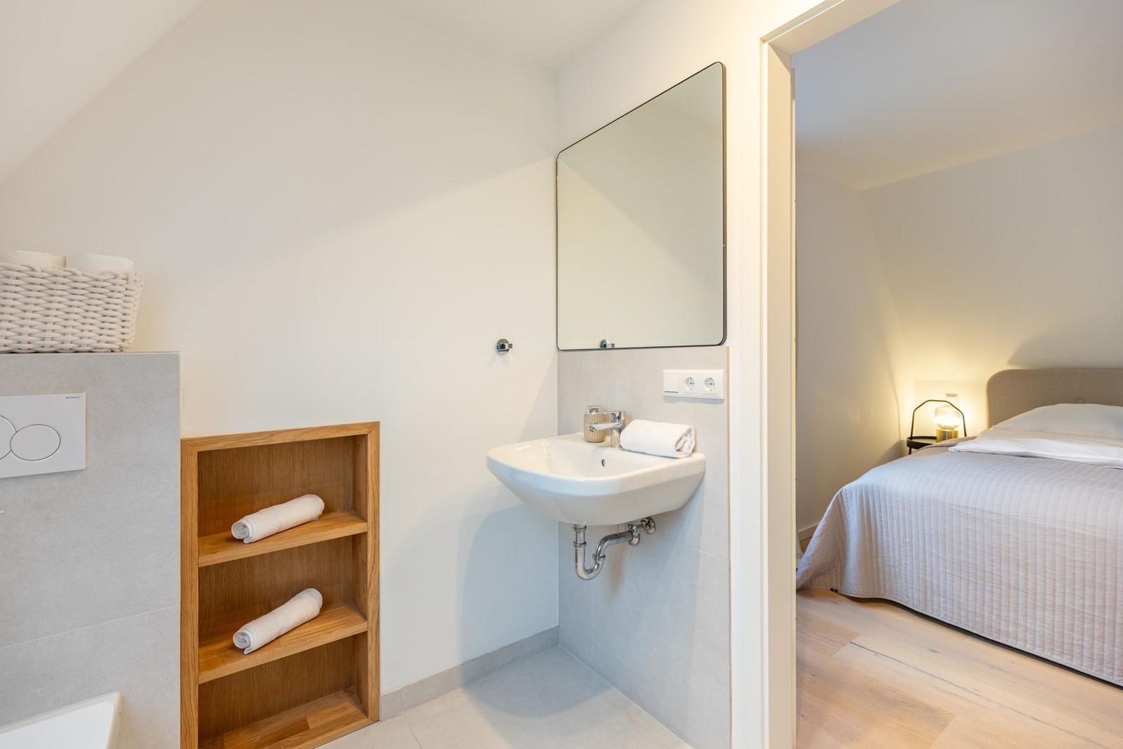 Badezimmer mit Waschbecken und Spiegel. Schlafzimmer mit Bett und Nachttisch sichtbar.