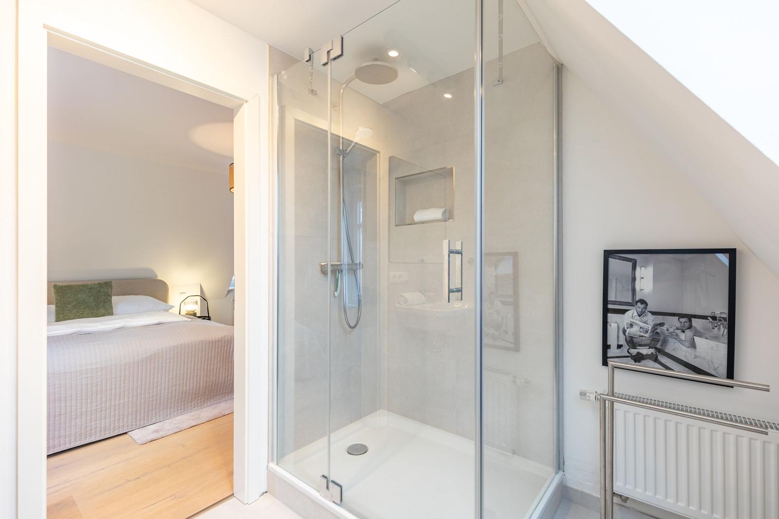 Duschbad mit Glaswand, Bett im Hintergrund, Wandbild und Heizkörper.