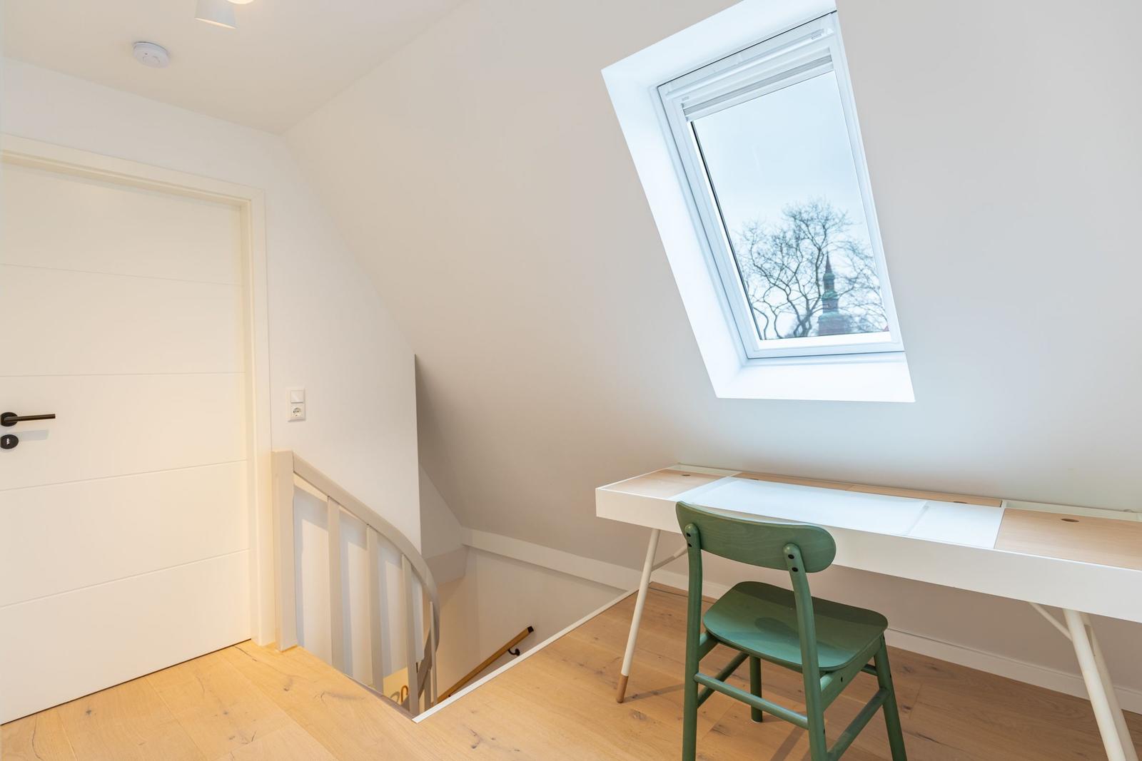 Dachgeschosszimmer mit Schreibtisch, grüner Stuhl und Dachfenster. Treppenhaus und Tür sichtbar.