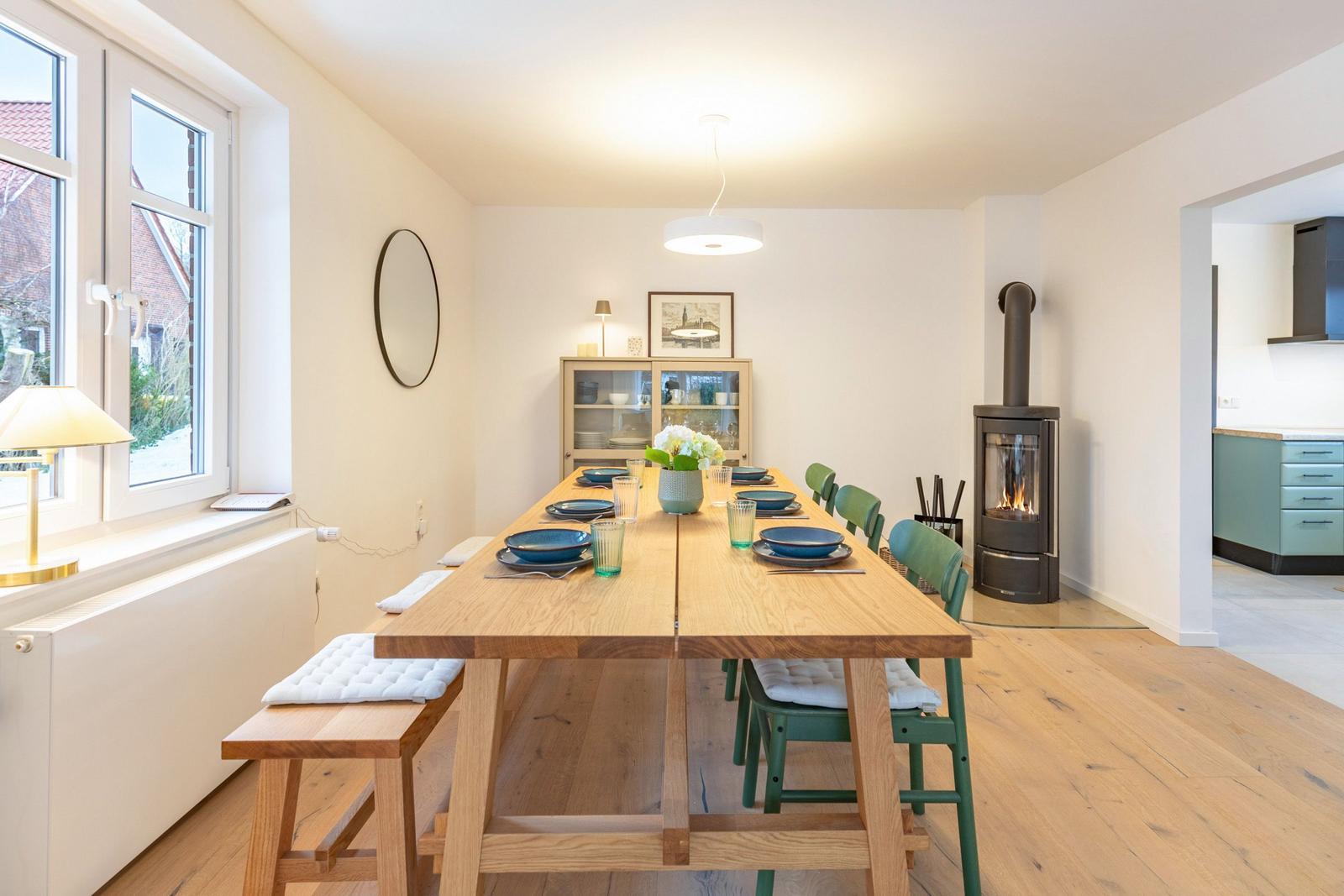 Esszimmer mit Holztisch, grünen Stühlen und Kaminofen. Fenster und Küche sind sichtbar.