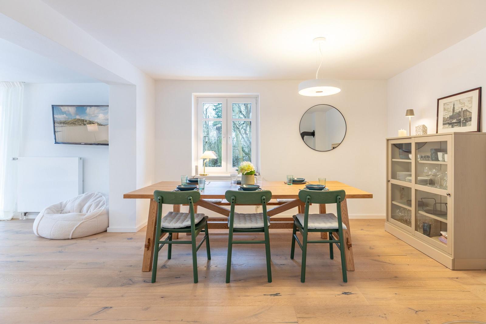 Essbereich mit Holztisch, grünen Stühlen und Fenster. Glaswand-Schrank und runder Spiegel an der Wand.