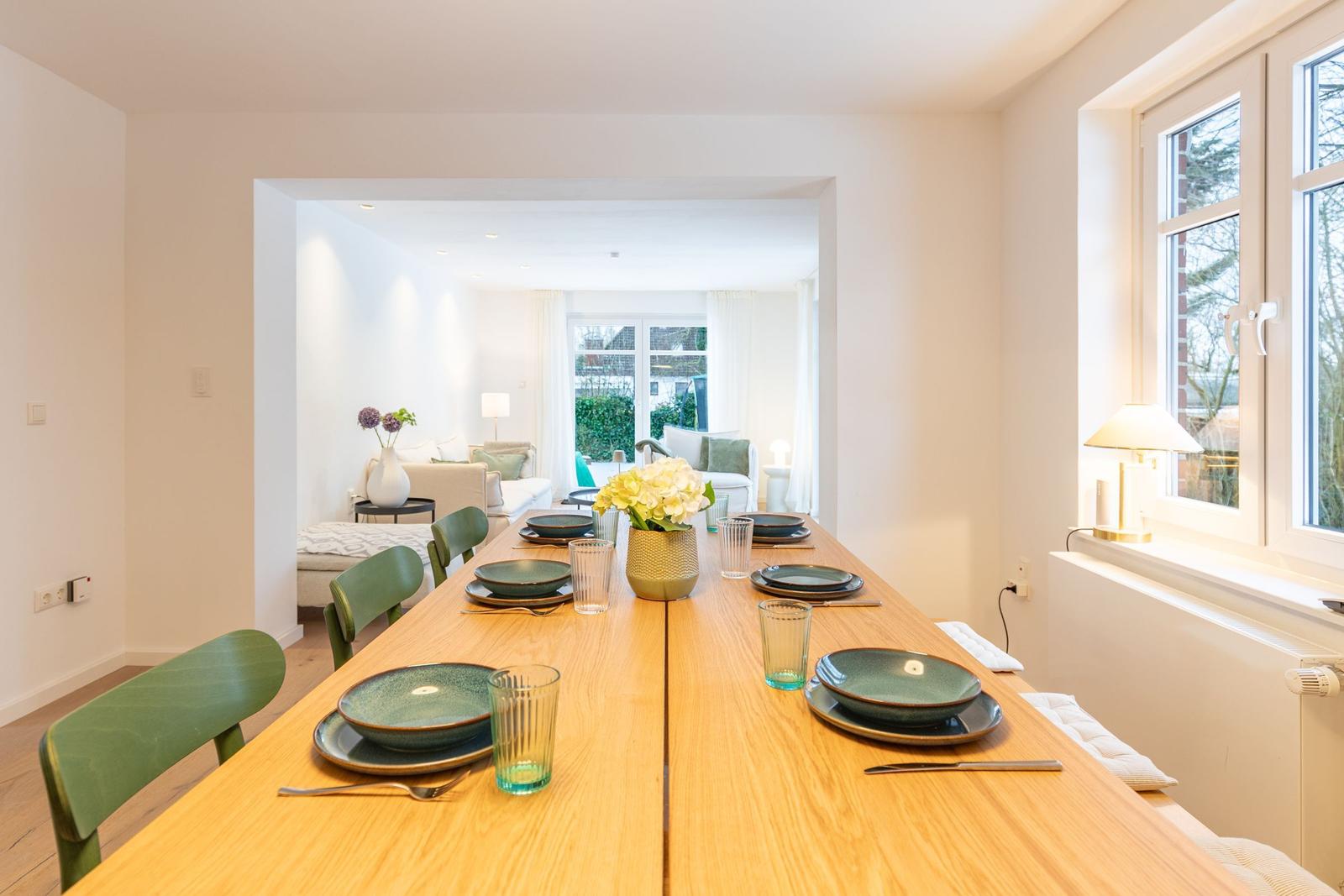 Esszimmer mit Holztisch und grünen Stühlen. Durchgang zum Wohnzimmer mit Sofa und Fenster.