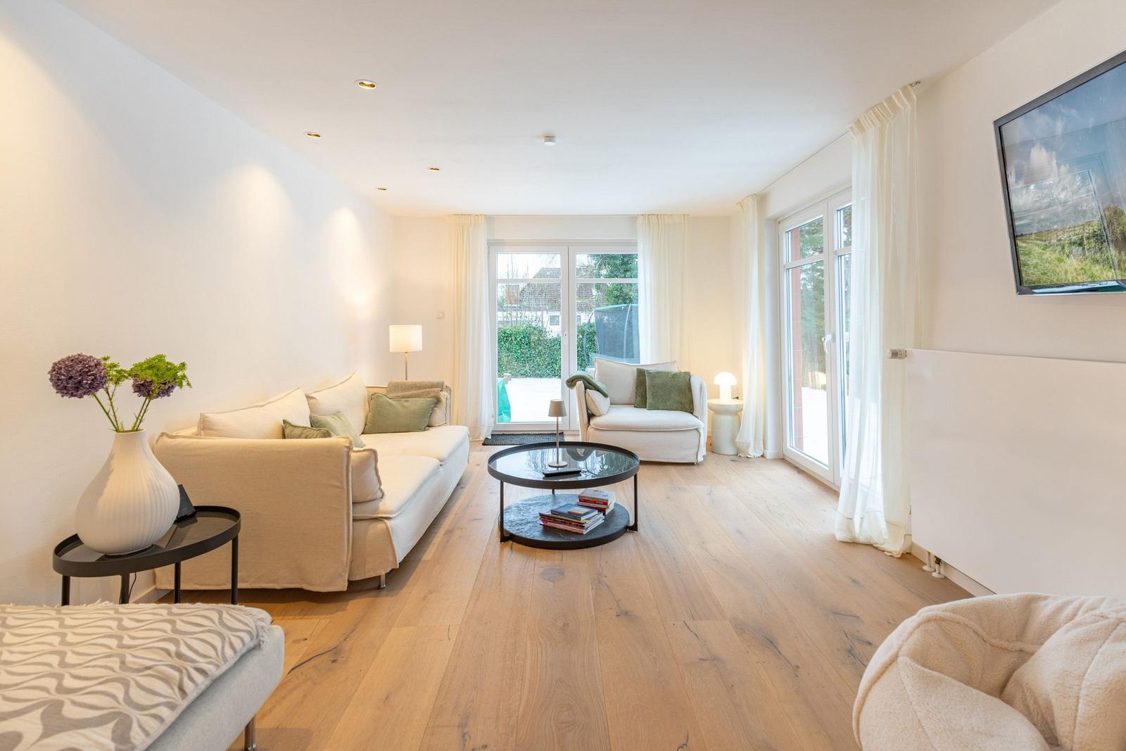 Wohnzimmer mit Sofa, Sessel und Glas-Tisch. Holzboden und große Fenster.