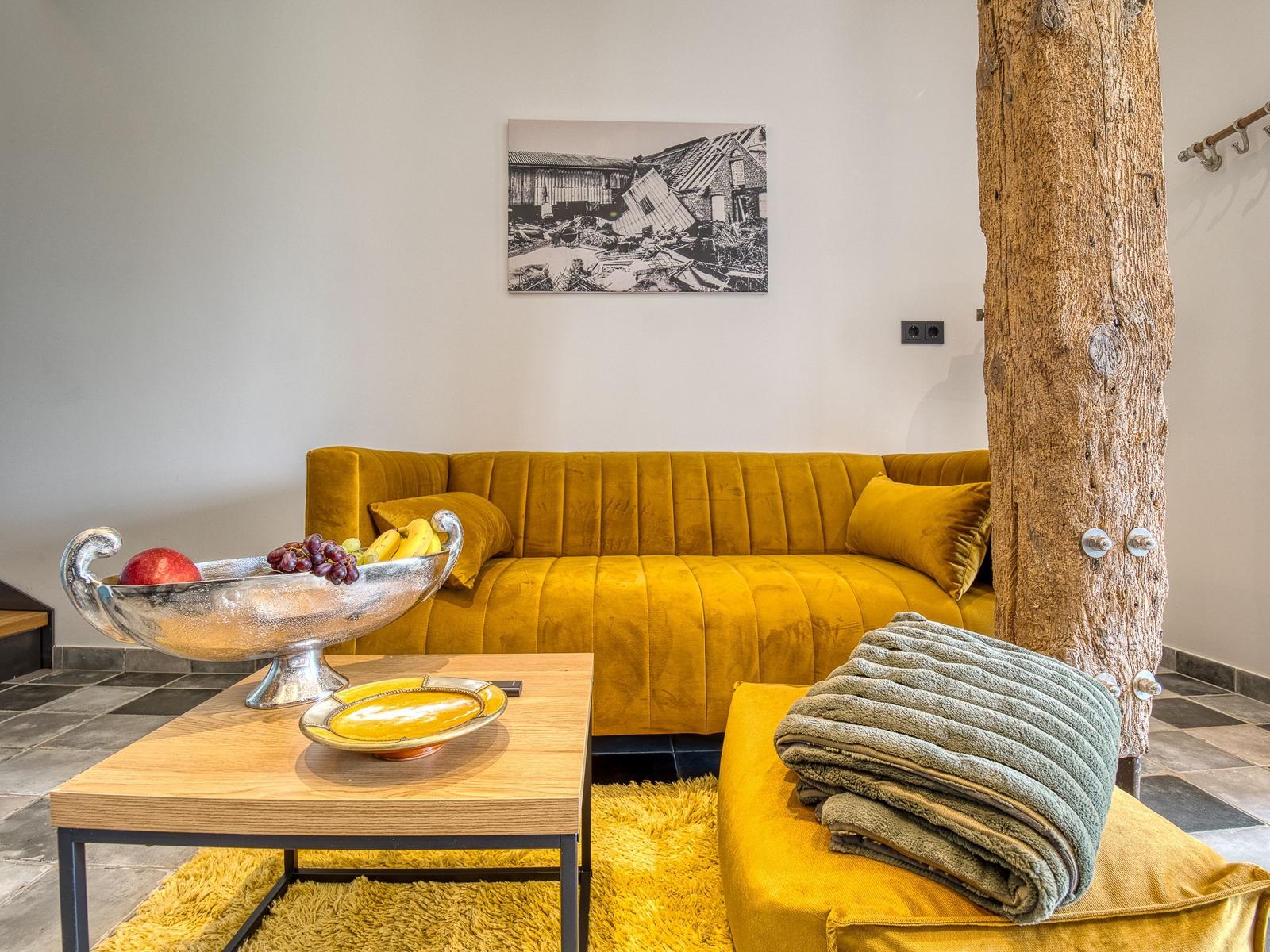 Gelber Sofa mit Kissen, Holztisch, Obstschale und großer Holzstütze.