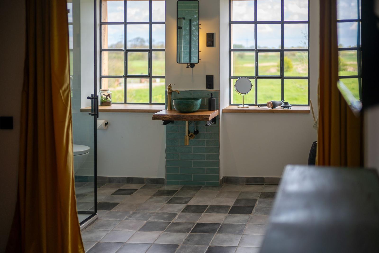 Badezimmer mit grünem Waschbecken, Holzunterschrank und großen Fenstern mit Blick auf Grünland.