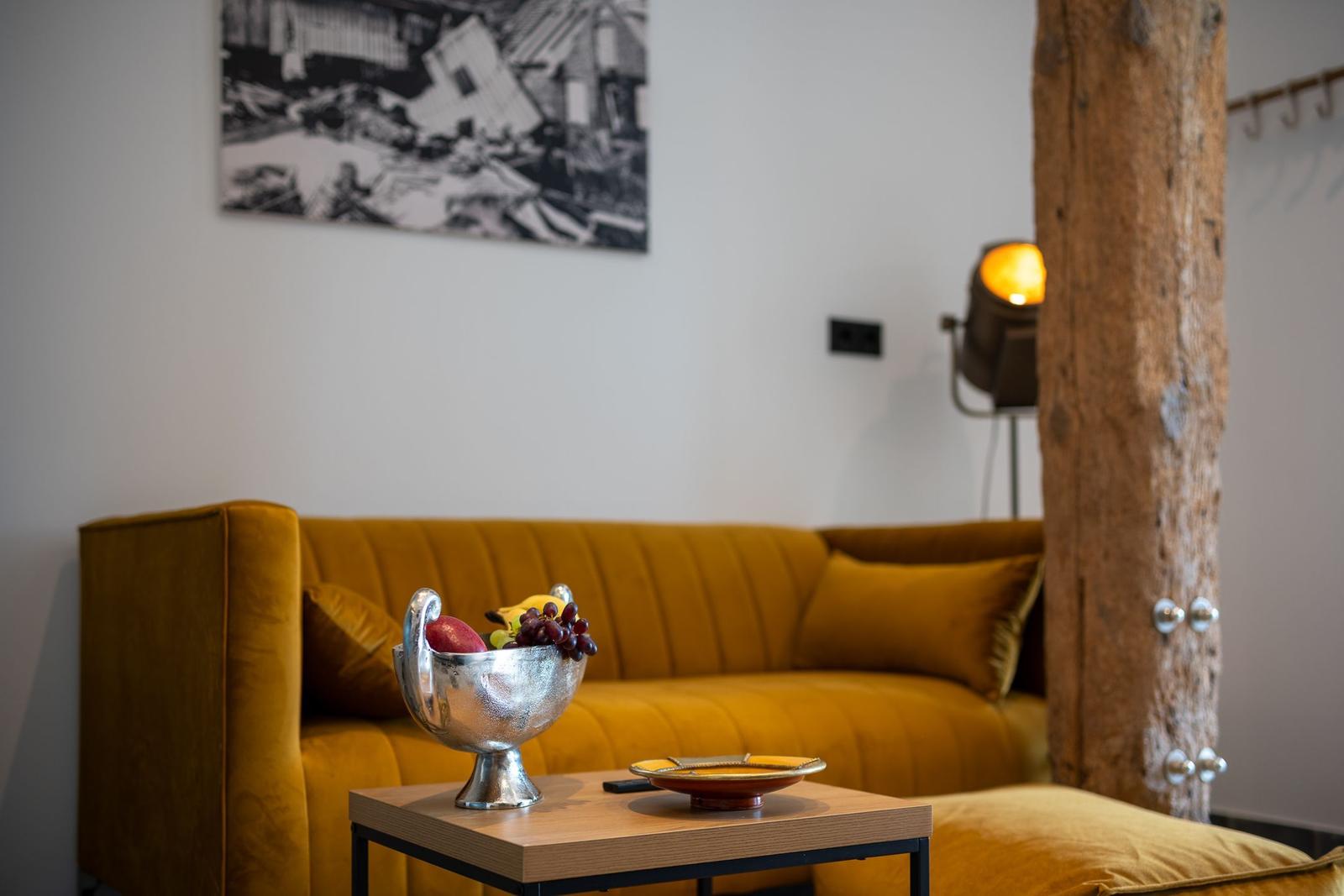 Gelber Sofa mit Obstschale auf Tisch, Holzpfosten und Wandbild im Hintergrund.