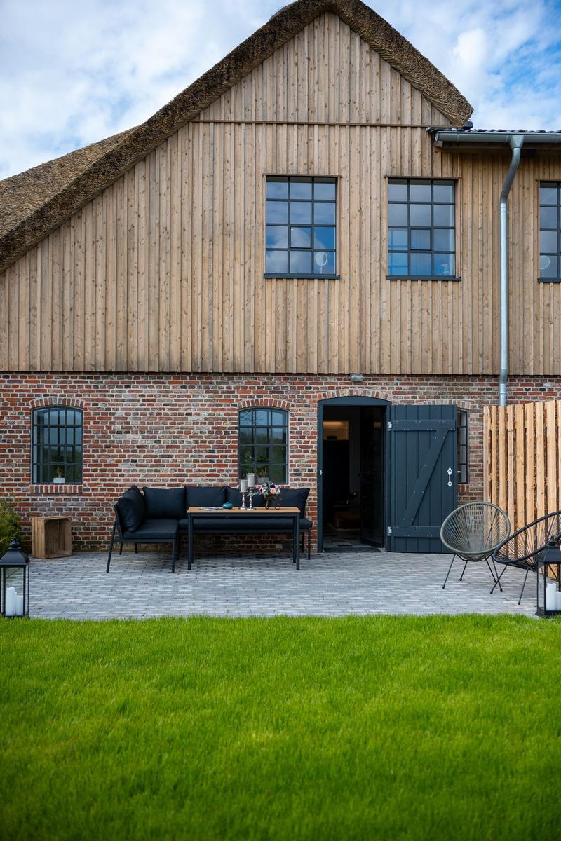 Haus mit Holzfassade und Dachstuhl, Terrasse mit Sitzgelegenheiten und Grasfläche.