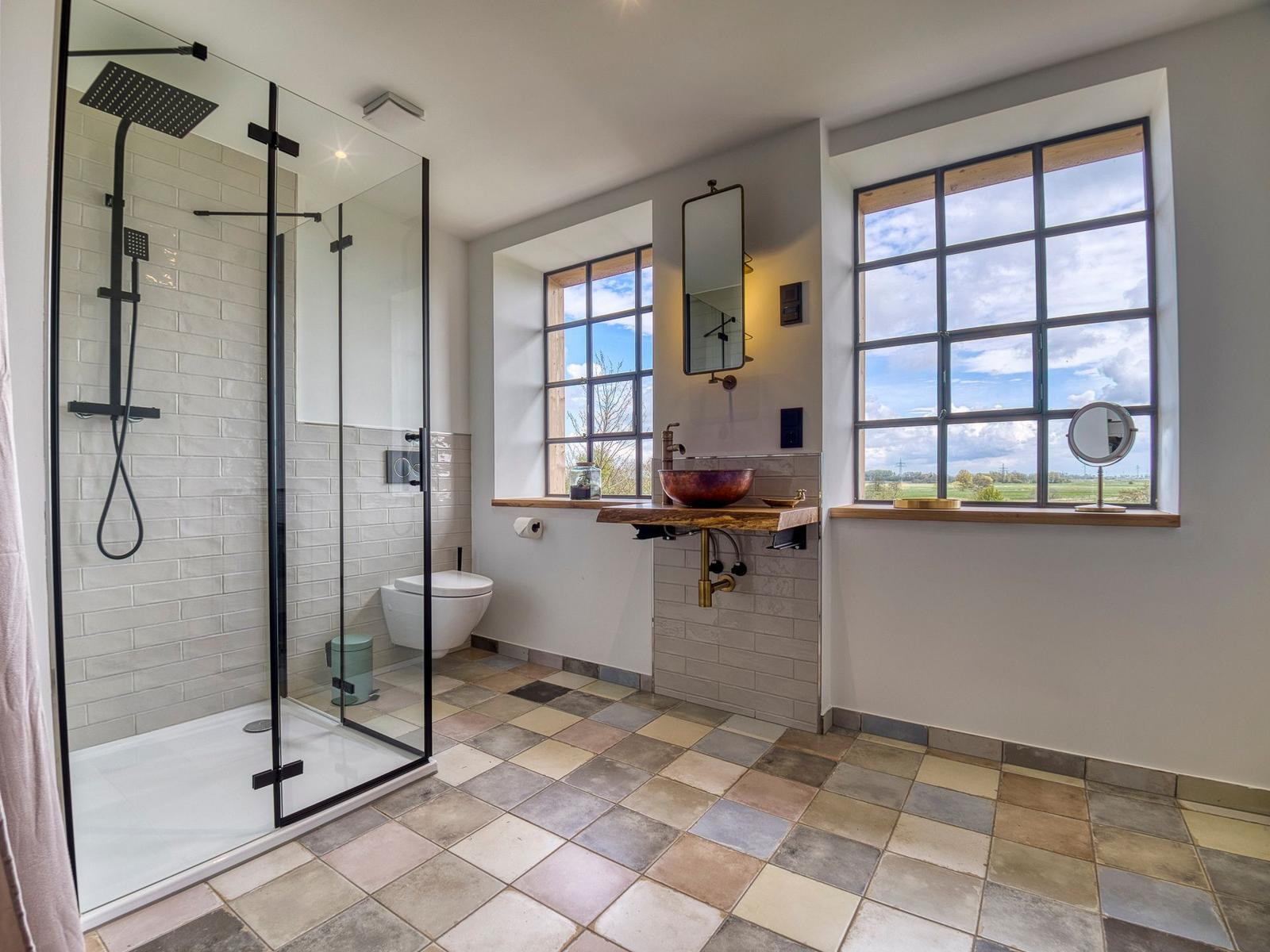 Badezimmer mit Dusche, Waschbecken und Fenstern mit Blick auf Landschaft.