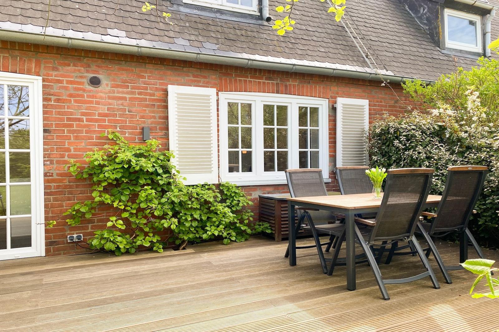 Terrasse mit Holztisch und Stühlen vor rotem Backsteinhaus mit weißen Fenstern.